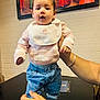 baby, child, indoor, person, hands, sweater, pants, table, painting, brick_wall, curious, expression, blue_jeans, sock, holding, portrait, cute, casual_clothing, watch, bracelet