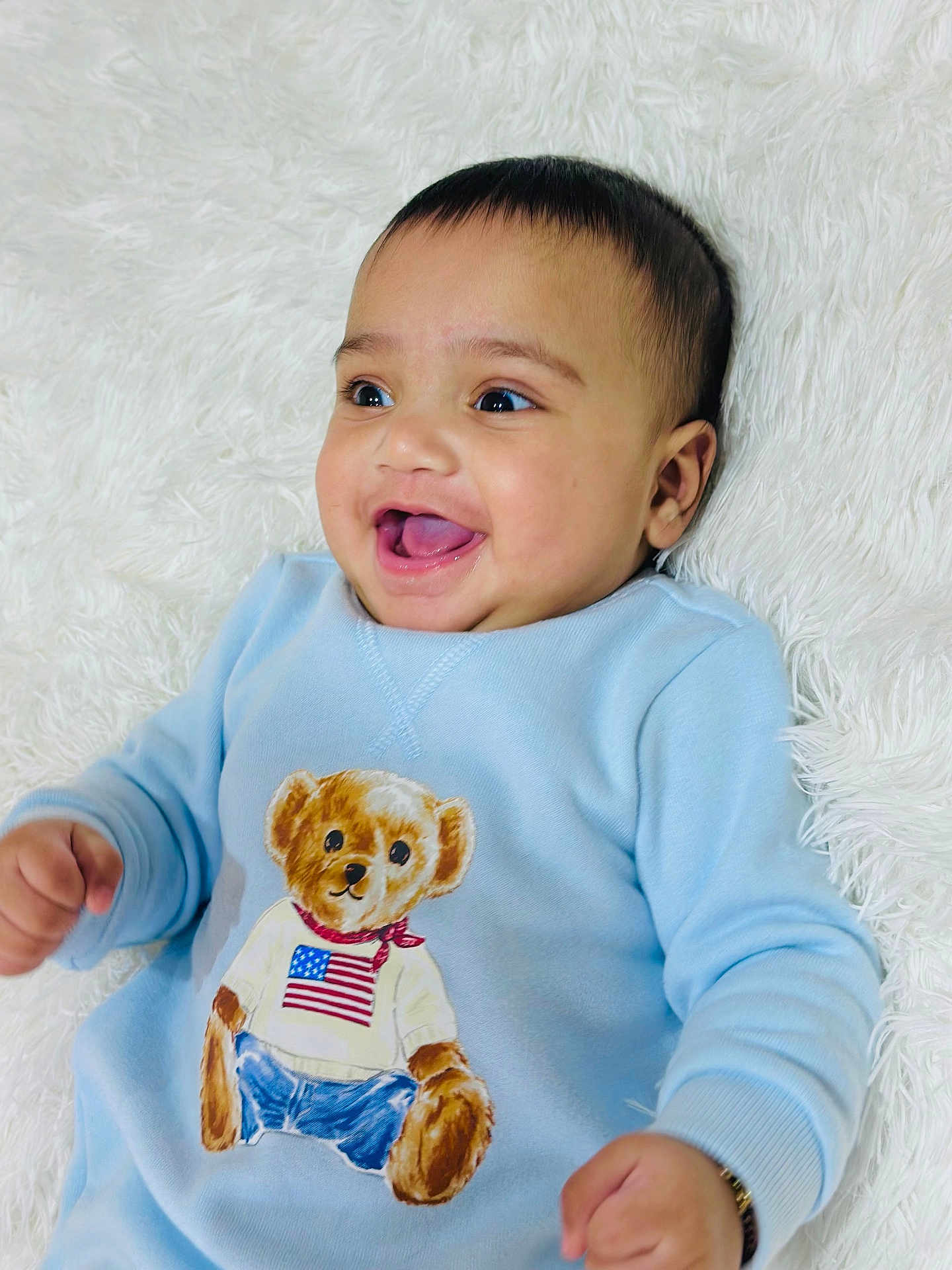 Bramh is registered to the contest to win money with this photo: baby, child, smiling, blue_sweater, teddy_bear, american_flag, infant, happy, lying_down, blanket, cute, portrait, face, clothing, person, young_child, indoors, bright_eyes, open_mouth, soft_texture