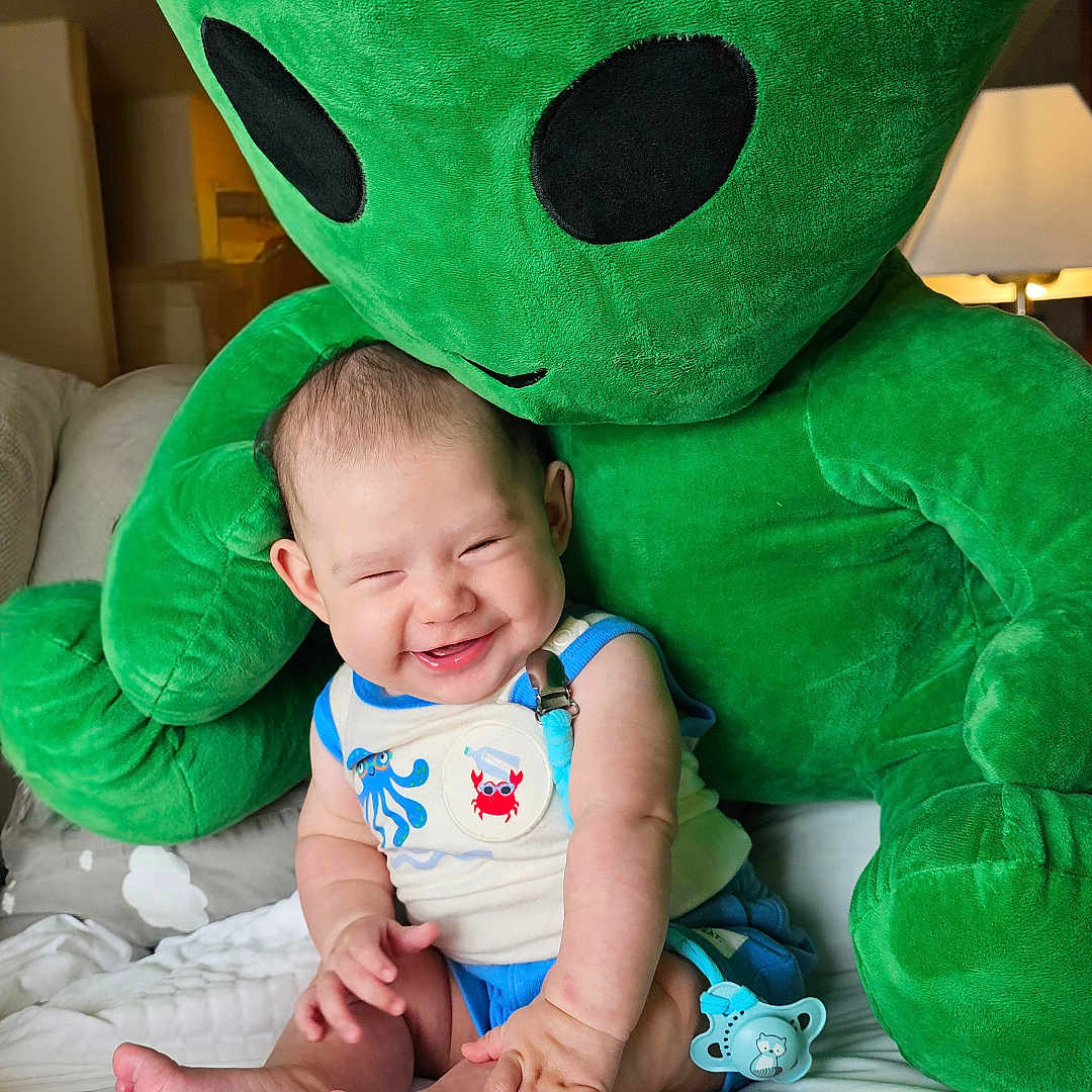 Leonora joined the competition — help win amazing prizes! baby, bed, child, childrens_toy, clothing, couch, cute, cute_outfit, green_alien, happy, home, indoor, infant, pacifier_clip, person, plush_toy, sitting, smiling, soft_toy, toy