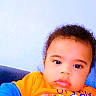 toddler, child, baby, face, portrait, blue_clothing, orange_clothing, indoor, couch, expression, curly_hair, skin, person, young_child, casual_wear, looking_at_camera, sitting, closeup, text_overlay, emoji