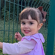 Victoria participe au concours pour gagner de l'argent avec cette photo : child, toddler, smiling, girl, outdoor, green_fence, brown_hair, bow, long_sleeve, grass, nature, portrait, happy, face, person, cute, young_child, daylight, casual_clothing, playful