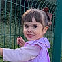 child, toddler, smiling, girl, outdoor, green_fence, brown_hair, bow, long_sleeve, grass, nature, portrait, happy, face, person, cute, young_child, daylight, casual_clothing, playful