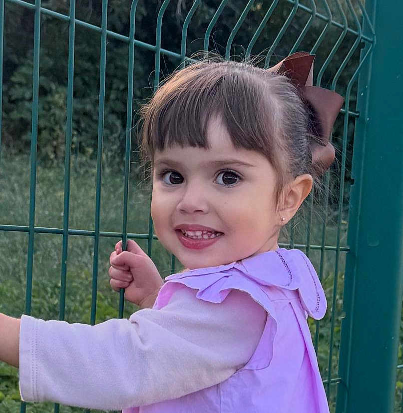 Victoria participe au concours pour gagner de l'argent avec cette photo : child, toddler, smiling, girl, outdoor, green_fence, brown_hair, bow, long_sleeve, grass, nature, portrait, happy, face, person, cute, young_child, daylight, casual_clothing, playful
