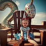 child, toddler, birthday, party_hat, balloon, blue_dress, barefoot, chair, wooden_furniture, outdoor, beach, sunny, celebration, happy, sunglasses, party, smiling, holiday, sky, festive