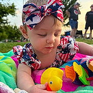 Mikayla is registered to the contest to win money with this photo: baby, child, rubber_duck, headband, outdoor, grass, blanket, toy, colorful, person, patriotic, red_white_blue, playing, cute, summer, people, blurred_background, infant, closeup, nature