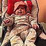 baby, infant, pajamas, hat, baby_seat, pillow, red_pillow, dinosaur_pattern, cozy, indoor, child, smiling, cushion, seatbelt, clothing, person, portrait, relaxing, warm_lighting, cute