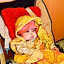 baby, blanket, hat, chair, cushion, red_pillow, indoors, child, warm_clothing, seated, face, portrait, cozy, fabric, textile, colorful, person, infant, wrapped, expression
