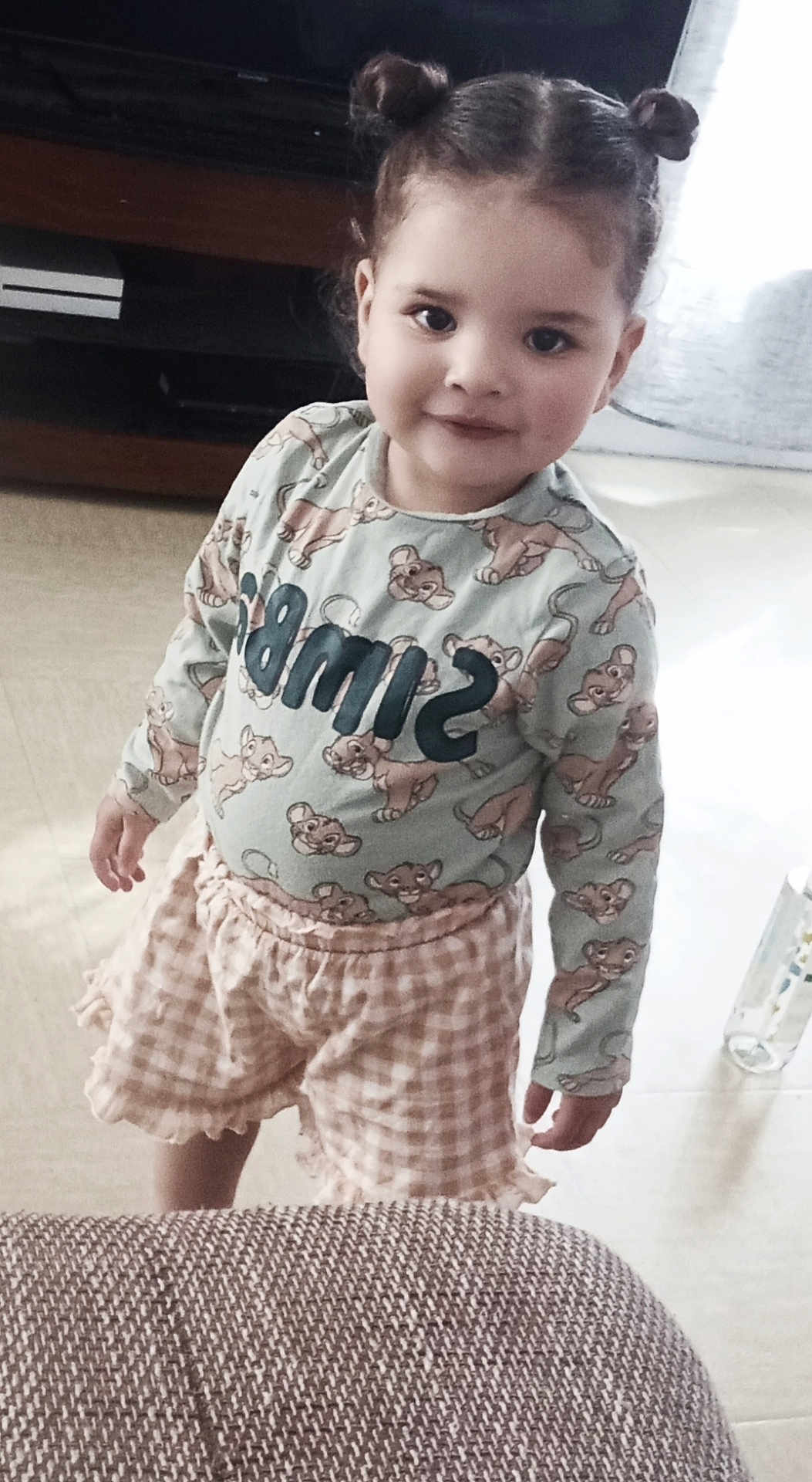Mia participe au concours pour gagner de l'argent avec cette photo : child, smiling, indoor, person, clothing, hair_buns, shorts, long_sleeve_shirt, floor, couch, bottle, cute, standing, young_child, portrait, playful, happy, home, furniture, light