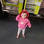child, toddler, firefighter_hat, pink_sweatshirt, fairy_design, barefoot, floor, fire_truck, yellow_vehicle, vehicle_steps, face, curious_expression, standing, indoors, casual_clothing, small_person, big_eyes, messy_face, red_hat, playful