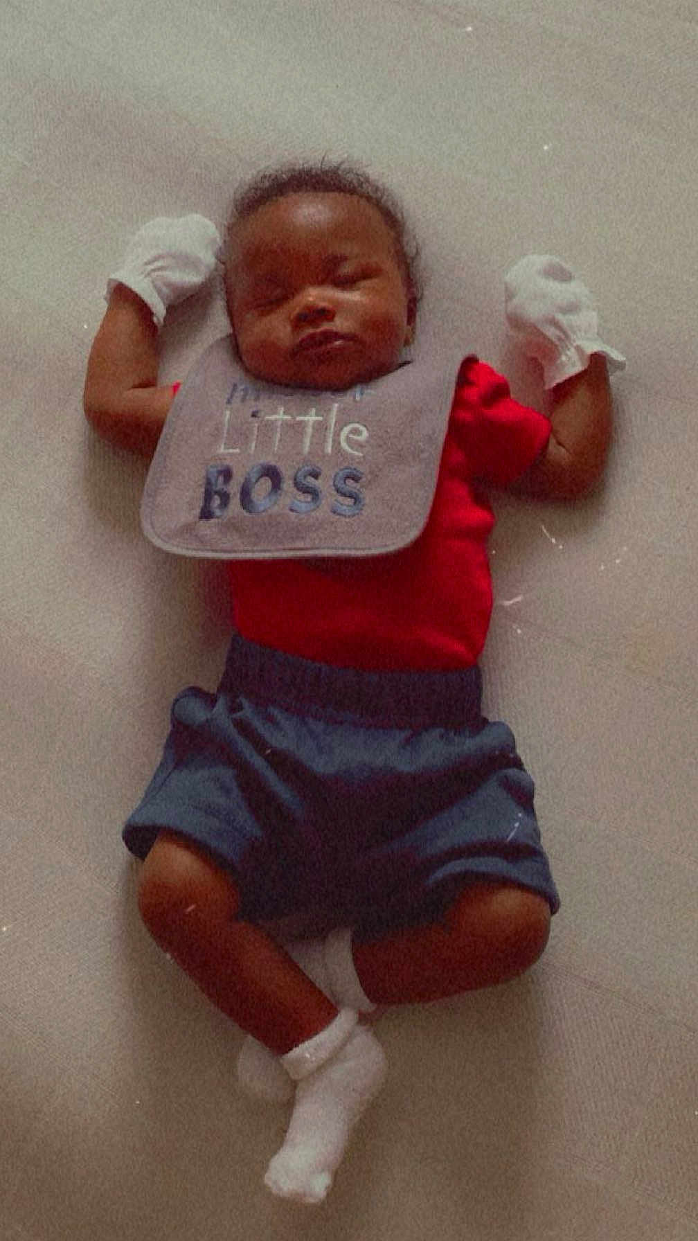 Skylan is registered to the contest to win money with this photo: baby, sleeping, infant, clothing, bib, red_shirt, blue_shorts, white_socks, mittens, lying_down, cute, peaceful, indoor, portrait, child, newborn, soft_surface, small_person, resting, adorable
