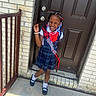 child, girl, smiling, dress, plaid, sash, grad, door, beads, hair, bracelet, white_shirt, black_shoes, socks, outdoor, concrete, railing, brick_wall, happy, portrait