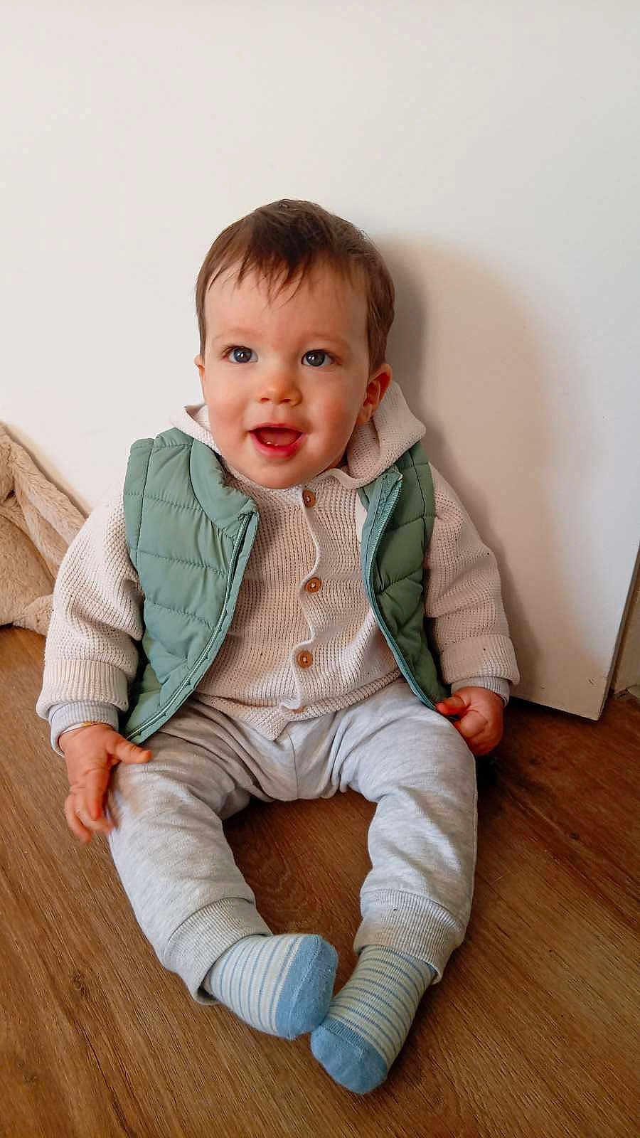 Lyzio participe au concours pour gagner de l'argent avec cette photo : baby, toddler, child, smiling, sitting, indoor, wooden_floor, green_vest, knit_sweater, buttons, striped_socks, pants, face, eyes, hands, legs, portrait, white_wall, cute, happy