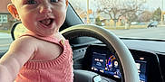 Mia is registered to the contest to win money with this photo: arm, baby, blue_shorts, car_interior, child, clothing, dashboard, daylight, face, happy, person, pink_top, portrait, road, seat, smiling, steering_wheel, trees, vehicle, window