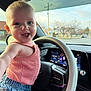 Mia is registered to the contest to win money with this photo: baby, child, steering_wheel, car_interior, pink_top, blue_shorts, smiling, happy, face, arm, window, dashboard, seat, road, trees, daylight, clothing, person, portrait, vehicle