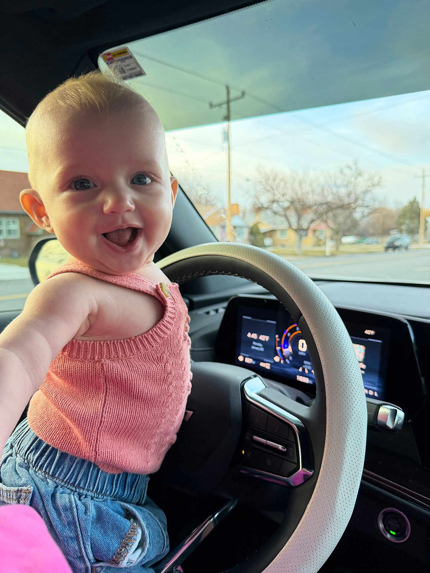 Mia is registered to the contest to win money with this photo: baby, child, steering_wheel, car_interior, pink_top, blue_shorts, smiling, happy, face, arm, window, dashboard, seat, road, trees, daylight, clothing, person, portrait, vehicle