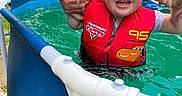 Isabella is registered to the contest to win money with this photo: adult, blue_pool, child, fun, greenery, hands, happy, life_jacket, outdoor, person, pool, red, safety, smiling, splash, summer, swimming, toddler, trailer, water