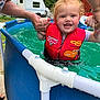 toddler, child, pool, water, life_jacket, red, smiling, outdoor, summer, hands, adult, safety, blue_pool, swimming, happy, greenery, trailer, splash, person, fun