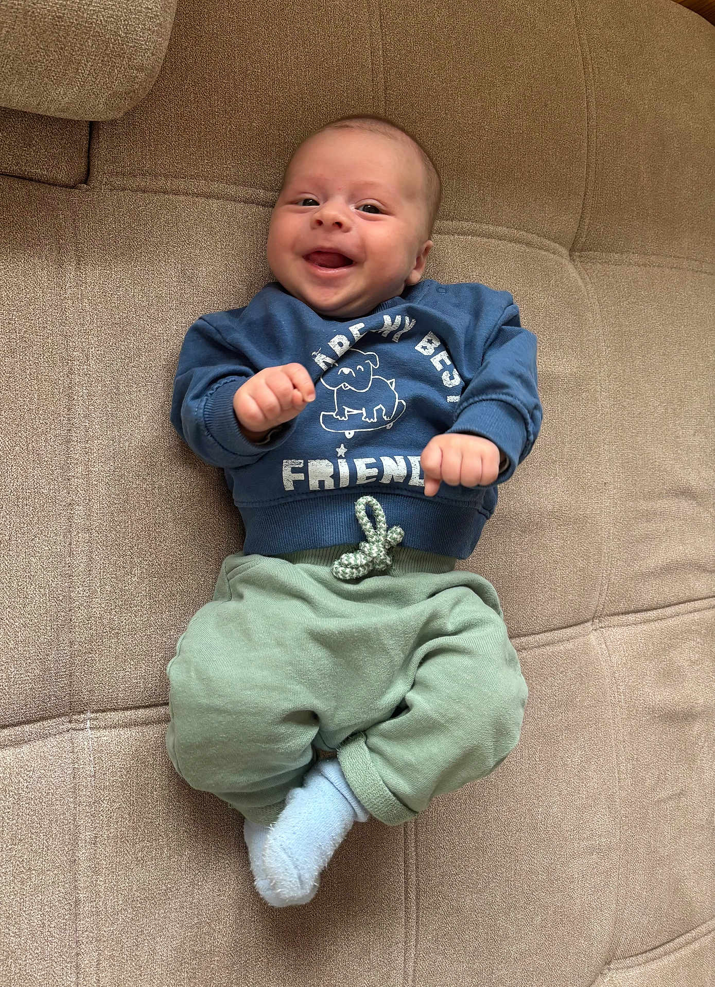 Jules participe au concours pour gagner de l'argent avec cette photo : baby, infant, smiling, blue_sweatshirt, green_pants, sock, couch, furniture, happy, face, person, child, clothing, indoors, cute, playful, young, sitting, cozy, portrait