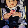 Neda is registered to the contest to win money with this photo: baby, infant, child, car_seat,  자동차, harness, buckled, sitting, face, eyes, hands, clothing, white_shirt, pants, portrait, indoors, vehicle, safety, wide_eyed, adorable