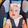 baby, child, infant, portrait, car_seat, safety_harness, pink_bow, headband, sitting, indoors, wide_eyed, curious, looking_at_camera, smiling, baby_clothing, pink_outfit, chair, seat_belt, close_up, safe