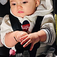 Neda is registered to the contest to win money with this photo: baby, infant, child, car_seat, seatbelt, buckled, portrait, face, hands, clothing, white_outfit, hoodie, sleeves, bracelet, indoor, vehicle, passenger, serious_expression, sitting, photograph