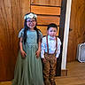 child, girl, boy, flower_crown, dress, bow_tie, suspenders, glasses, wooden_door, wooden_floor, indoor, smile, serious, standing, siblings, portrait, clothing, footwear, home, fan