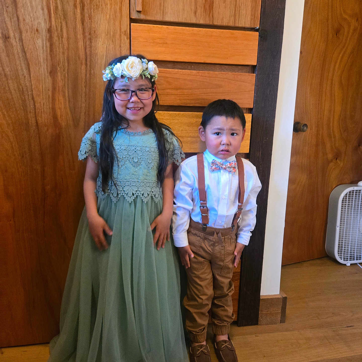 Elia is registered to the contest to win money with this photo: child, girl, boy, flower_crown, dress, bow_tie, suspenders, glasses, wooden_door, wooden_floor, indoor, smile, serious, standing, siblings, portrait, clothing, footwear, home, fan