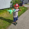 child, boy, firefighter_hat, toy, plush_dinosaur, outdoor, grass, sidewalk, house, car, sunny, shadow, playful, casual, summer_clothing, greenery, residential_area, smiling, daytime, fun