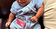 Ace is registered to the contest to win money with this photo: baby, child, smiling, blue_clothing, romper, cartoon_print, leather_couch, pillow, sitting, sandals, happy, cute, portrait, indoors, infant, face, skin, footwear, shorts, hand