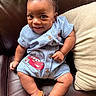 baby, child, smiling, blue_clothing, romper, cartoon_print, leather_couch, pillow, sitting, sandals, happy, cute, portrait, indoors, infant, face, skin, footwear, shorts, hand