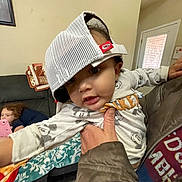 Skyler is registered to the contest to win money with this photo: baby, child, toddler, cap, mesh_cap, hat, onesie, pajamas, couch, sofa, blanket, pillow, tablet, reclining_child, adult_hand, living_room, door, window, portrait, smile