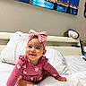 baby, child, infant, smiling, sitting, portrait, pink_bow, headband, pink_clothing, dress, hearts, bed, white_bedding, pillow, stuffed_toy, bedroom, wall_art, cityscape, bridge, indoor