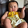 baby, child, yellow_dress, sitting, leather_chair, indoor, curious_expression, skin, face, arms, legs, buttoned_clothing, patterned_skirt, black_hair, infant, portrait, young_child, furniture, cute, person
