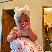 Delilah is registered to the contest to win money with this photo: baby, child, indoor, dress, headband, white, red, hand, pointing, smiling, person, room, door, closet, clothing, furniture, portrait, cute, happy, infant