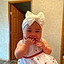 Delilah is registered to the contest to win money with this photo: baby, child, indoor, dress, headband, bow, fingers, smiling, cute, infant, wooden_door, floor, wall, person, portrait, happy, sleeveless, room, skin, hand