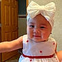 Delilah is registered to the contest to win money with this photo: baby, child, white_dress, red_flower_embroidery, headband, white_bow, smiling, indoors, hand_holding, wooden_door, beige_wall, cute, portrait, person, infant, adorable, happy, face, clothing, home