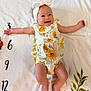 Briella is registered to the contest to win money with this photo: baby, infant, smiling, headband, flower_pattern, romper, blanket, floral, numbers, arms_outstretched, cute, happy, lying_down, indoors, skin, person, child, head, feet, toddler
