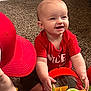Nora is registered to the contest to win money with this photo: baby, infant, child, smile, red_shirt, red_hat, toy_blocks, plastic_toy, carpet, sitting, happy, hands, eyes, face, indoor, playtime, cheeks, clothing, portrait, cute