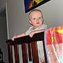 baby, infant, crib, wooden_rail, blanket, colorful_blanket, blue_eyes, standing, hands, toys, mattress, painting, wall_art, indoor, portrait, flash_photography, bedroom, feet, doorway, child