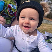 Nora is registered to the contest to win money with this photo: baby, infant, smiling, blue_eyes, beanie_hat, pompom, onesie, outdoor, grass, trees, portrait, close_up, cheeks, rosy_cheeks, parent_hand, colorful_tshirt, cartoon_print, safety_strap, happy, sunlight