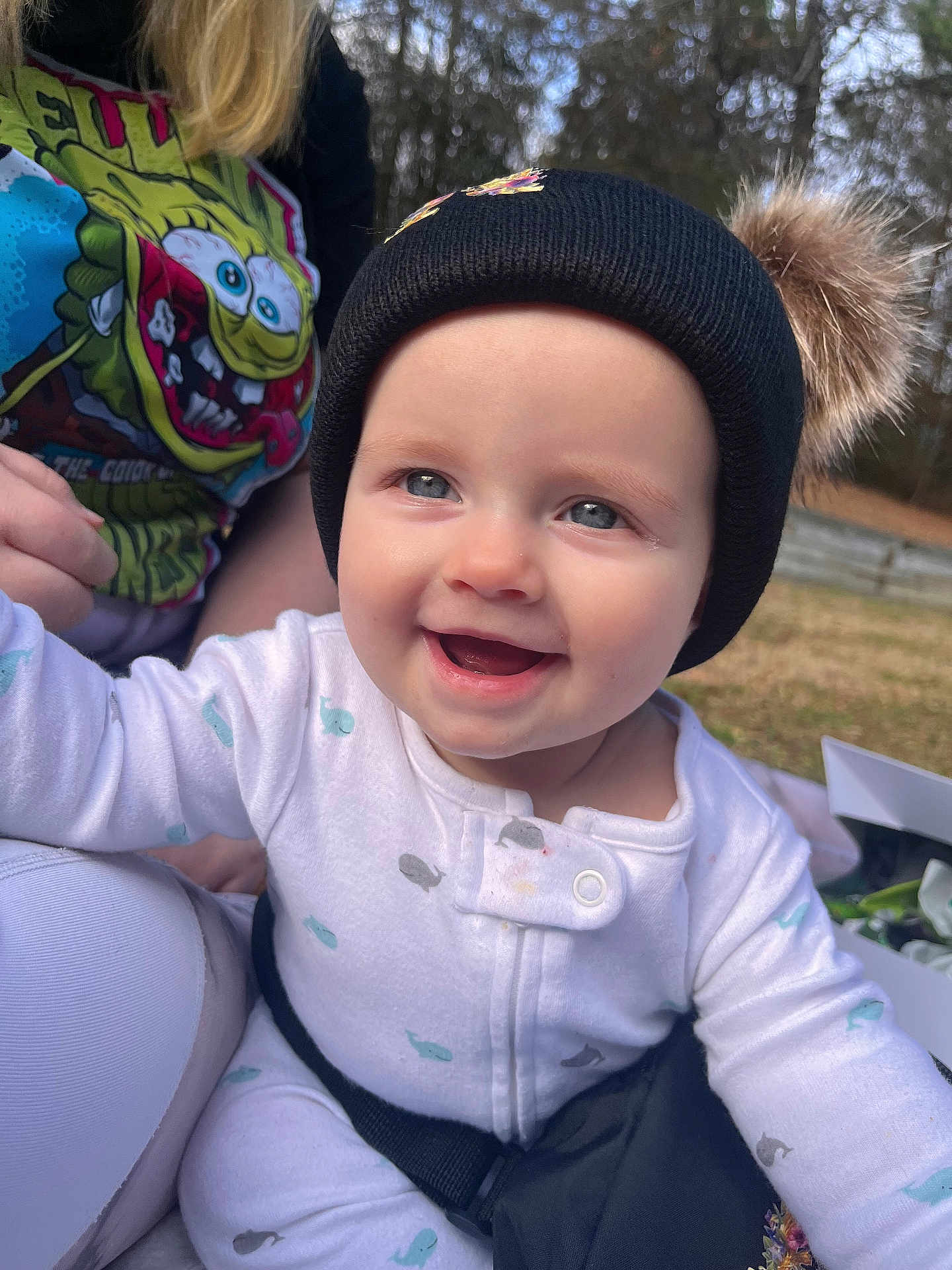 Nora is registered to the contest to win money with this photo: baby, infant, smiling, blue_eyes, beanie_hat, pompom, onesie, outdoor, grass, trees, portrait, close_up, cheeks, rosy_cheeks, parent_hand, colorful_tshirt, cartoon_print, safety_strap, happy, sunlight
