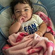 Ezaiah is registered to the contest to win money with this photo: baby, infant, child, portrait, smiling, happy, sitting, blanket, pink_blanket, baby_seat, blue_chair, indoors, face, dark_hair, brown_eyes, hand, finger_sucking, white_shirt, clothing, cozy