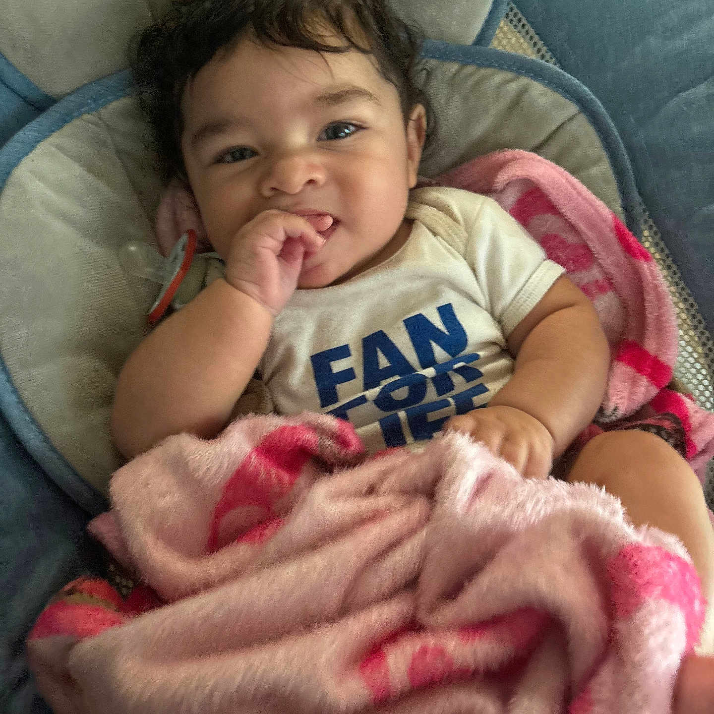Ezaiah is registered to the contest to win money with this photo: baby, infant, child, portrait, smiling, happy, sitting, blanket, pink_blanket, baby_seat, blue_chair, indoors, face, dark_hair, brown_eyes, hand, finger_sucking, white_shirt, clothing, cozy