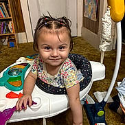 Juliana is registered to the contest to win money with this photo: toddler, child, baby, smiling, sitting, activity_walker, toy, playroom, indoor, portrait, face, hair_bows, floral_shirt, colorful, bookshelf, books, door, carpet, children_toys, happy