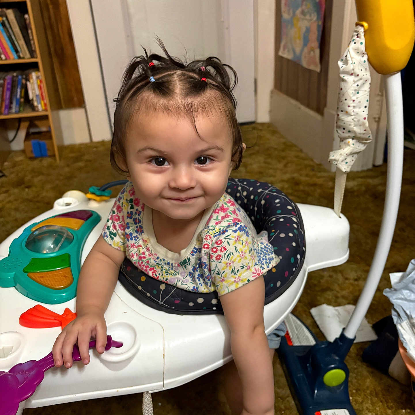 Juliana is registered to the contest to win money with this photo: toddler, child, baby, smiling, sitting, activity_walker, toy, playroom, indoor, portrait, face, hair_bows, floral_shirt, colorful, bookshelf, books, door, carpet, children_toys, happy
