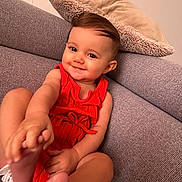 Inaya a rejoint le concours — aidez-le/la à gagner de superbes lots ! baby, child, smiling, red_clothing, couch, pillow, indoors, cute, happy, playful, skin, feet, hand, face, person, portrait, infant, sitting, cozy, home