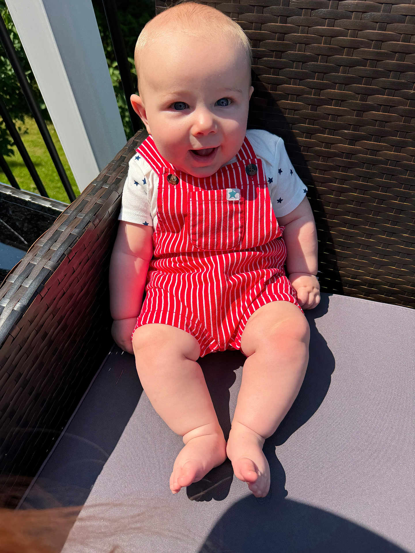 Parker is registered to the contest to win money with this photo: baby, child, smiling, outdoor, sunlight, red_overalls, white_shirt, star_pattern, chair, woven_chair, legs, feet, happy, cute, infant, summer, nature, greenery, portrait, seat