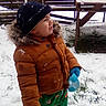 backyard, bench, black_hat, child, cold_weather, fur_hood, gloves, grass, green_pants, mittens, orange_jacket, playful, sippy_cup, smiling, snow, snowflakes, standing, toddler, winter_coat, wooden_fence