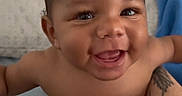Makari is registered to the contest to win money with this photo: baby, smile, happy, infant, tattoo, spider_web, arm, skin, diaper, curly_hair, face, person, indoor, bed, blanket, cute, sitting, child, expression, joy