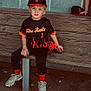 baseball, bench, black_uniform, cap, child, cleats, concrete_wall, dirt, face, fire_design, glove, hat, kid, outdoor, red_glove, sitting, sport_equipment, sports, team_uniform, young_player